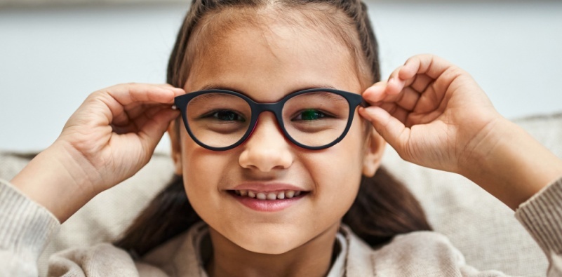 A young girl wearing black eyeglasses, holding on to each of the sides and smiling.