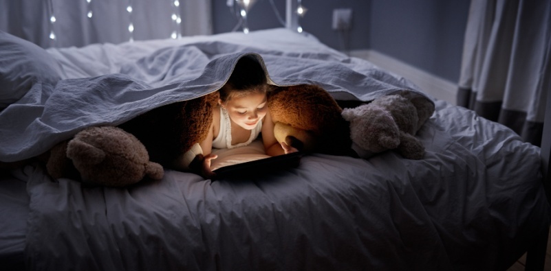 A young girl on her tablet under the covers at night.