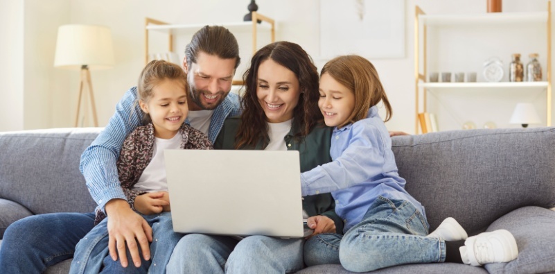 Smiling parents sitting on sofa with two children, hugging, using laptop together.