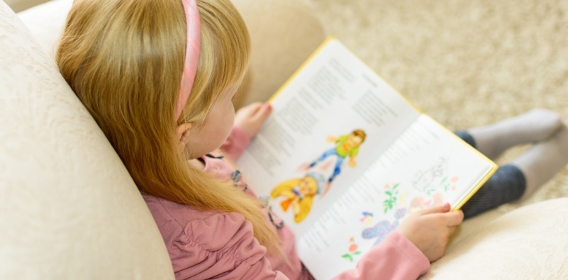 An over the shoulder view of a young girl sat on the couch reading a book.