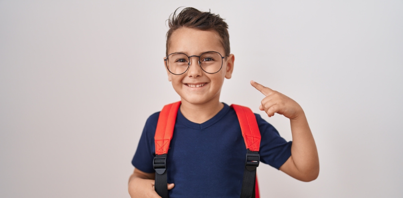 A young boy wearing a blue t-shirt and red backpack pointing to his round eyeglasses