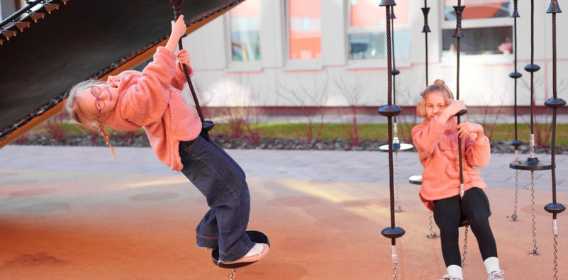 Two young girls wearing pink jackets and blue pants playing on the swings at a playground, the child on the left wears glasses.]
