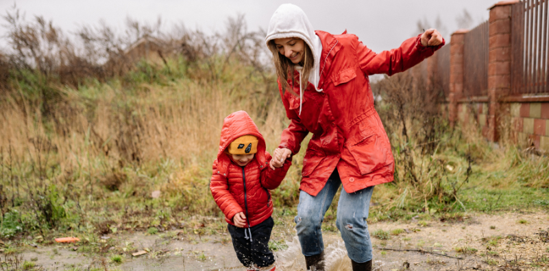 A young boy and his mum both wearing red coats, jumping in puddles outside.