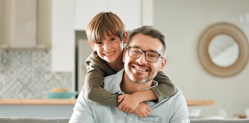 A young boy hugging his dad from behind. The dad is wearing a pair of spectacles.
