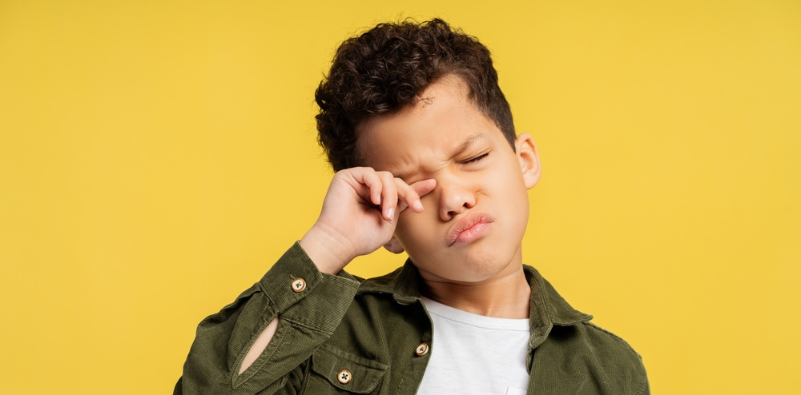 A young boy wearing a white t-shirt and green jacket, rubbing his right eye.