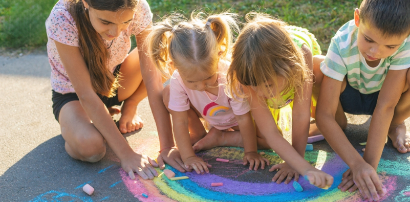 Four young children playing outside, drawing on the ground with colorful chalk.