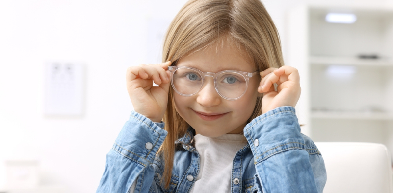 A young girl in a white t-shirt and denim jacket, holding the sides of her clear eyeglasses.