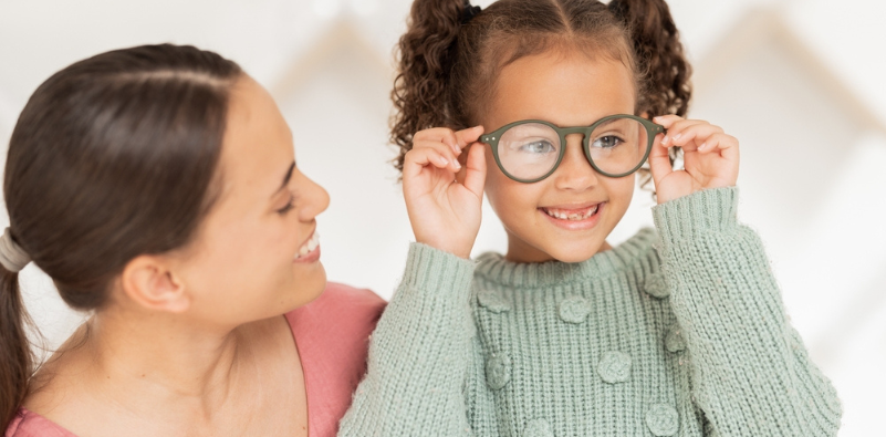 A child putting on her glasses, while her mum looks at her