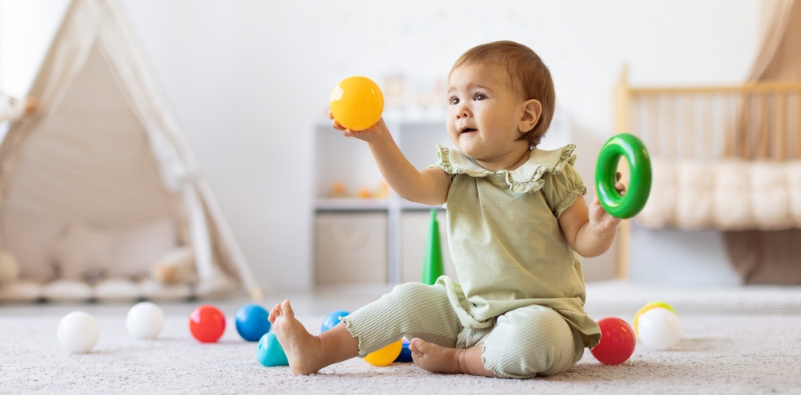 A smiling baby sitting on the floor holding a colorful ball and ring toy.