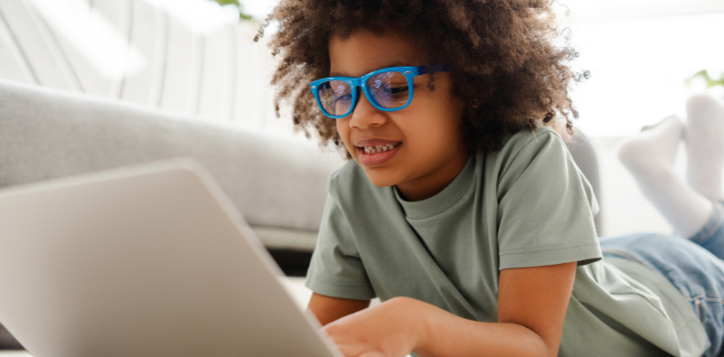 A young boy lying on the floor on a laptop, wearing a pair of blue eyeglasses