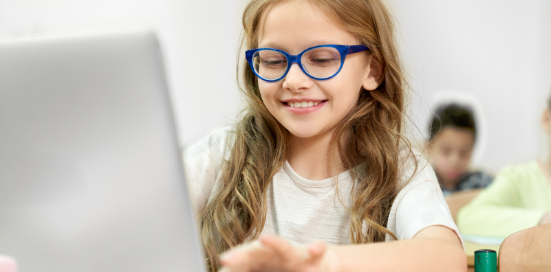 A young girl smiling and wearing glasses while using a laptop in school
