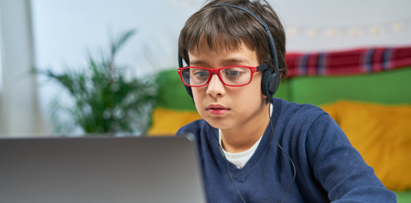 A child wearing red glasses and headphones while using a laptop.