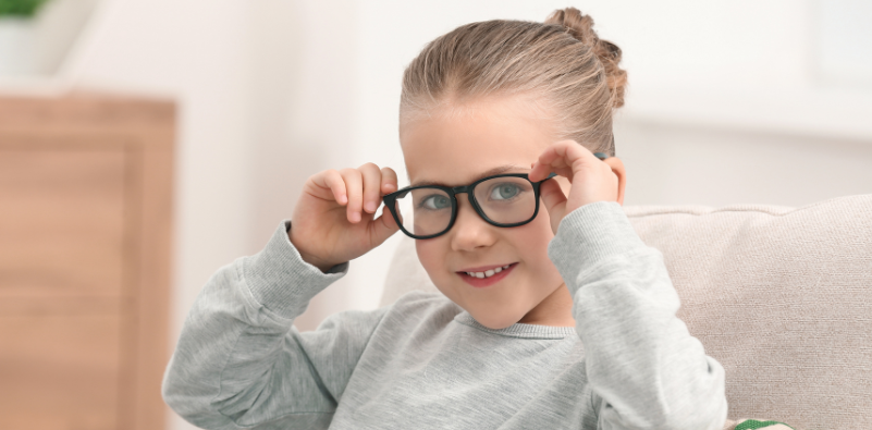 A young girl holding the sides of her black eyeglasses, while sat on the couch